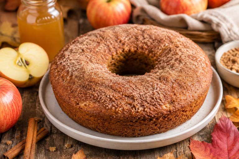 A high-quality, natural food photograph of a freshly baked Apple Cider Protein Donut Cake on a rustic wooden table. The cake is topped with a light cinnamon sugar coating and surrounded by fresh apples, cinnamon sticks, and autumn leaves. Bright, airy lighting with warm fall tones. Premium food photography style. No text overlay.